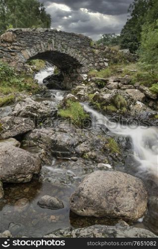Beautiful long exposure landscape image of Ashness Bridge in English Lake District during late Summer afternoon with moody weather