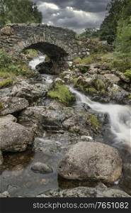 Beautiful long exposure landscape image of Ashness Bridge in English Lake District during late Summer afternoon with moody weather