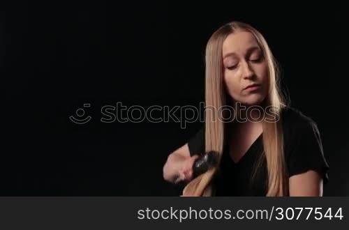Beautiful long hair blond woman combing her wonderful straight hair and looking with regret at the comb. Hair loss. Frustrated young female surprised she is losing hair. Black background. Human face expression emotion. Beauty hairstyle concept.