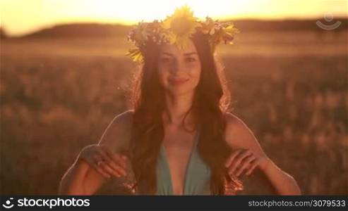 Beautiful long hair brunette woman in fashionable dress holding flower wreath over her head. Girl raises hands, taking off the wreath. Glow of setting sun shining through the wreath as she stands in golden wheat field in twilight time. Slow motion.