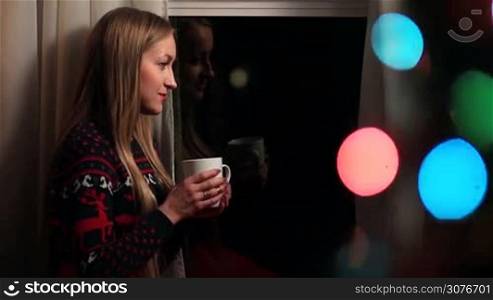 Beautiful long haired blonde woman standing by the window smiling and drinking fresh hot cup of coffee on Christmas eve. Foreground blurred colorful bokeh circles of christmas lights on tree.