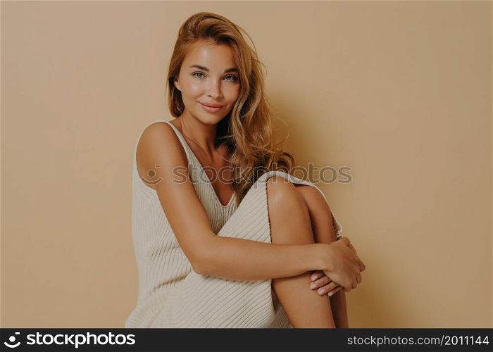 Beautiful long haired woman sits with crumpled up legs wears long dress shows bare knees and shoulders looks tenderly at camera isolated over beige background makes professional photo in studio. Beautiful long haired woman sits with crumpled up legs wears long dress isolated on beige background