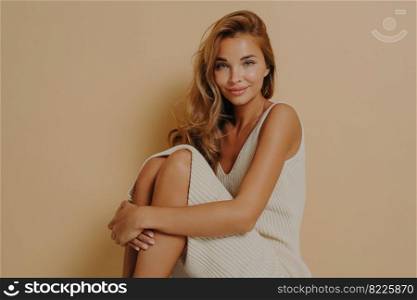 Beautiful long haired woman sits with crumpled up legs wears long dress shows bare knees and shoulders looks tenderly at camera isolated over beige background makes professional photo in studio. Beautiful long haired woman sits with crumpled up legs wears long dress isolated on beige background