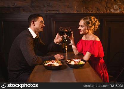 Beautiful love couple raised glasses with red wine in restaurant, romantic date. Elegant woman in red dress and her man, anniversary celebration