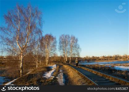 Beautiful March landscape with birches, a railway that stretches into the distance and a girl walking along the cross ties in the rays of the setting sun.