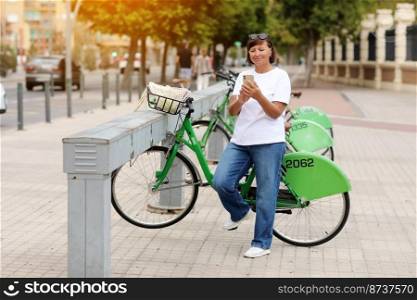 Beautiful mature woman holding smartphone and smiling while standing on the street near bicycles. Female with mobile phone in her hand using bike rental service on summer sunny day. Active lifestyle.. Beautiful mature woman holding smartphone and smiling while standing on the street near bicycles. Female with mobile phone in her hand using bike rental service on summer sunny day. Active lifestyle