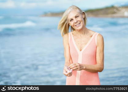 Beautiful mature woman walking along the shore of a tropical beach, wearing a nice orange dress. Elderly female enjoying her retirement at a seaside retreat.. Mature woman on shore of a beach. Elderly female enjoying her retirement at a seaside retreat.