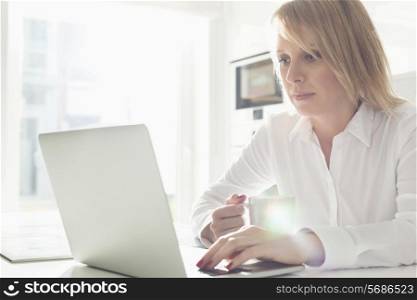 Beautiful mid adult businesswoman using laptop while having coffee at home