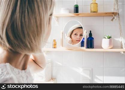 Beautiful middle-aged woman looking in the mirror in white eco friendly bathroom. Wooden shleves and reusable cosmetics bottles. Wellnes concept. Beautiful middle-aged woman looking in the mirror