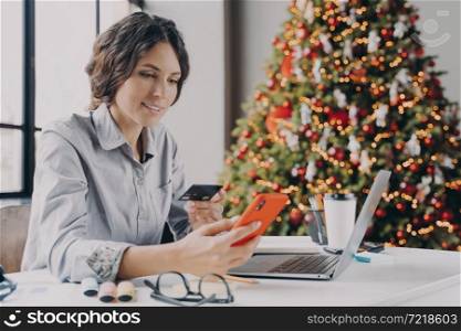 Beautiful millennial Spanish businesswoman holding credit card in hand and making payment on smartphone during Christmas holidays, buying gifts online while sitting at home office with Xmas tree. Spanish businesswoman holding credit card, doing online shopping on smartphone during Christmas time