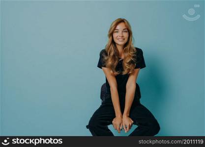 Beautiful millennial young lady with perfect styled blonde hair in casual black clothes sitting leaning with hands on edge of chair, bends forward and looks straight at camera with beaming smile. Beautiful young smiling lady hair in black casual t-shirt and jeans sitting on chair in studio