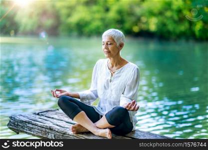 Beautiful mindful senior woman meditating by the water. Meditating by the water