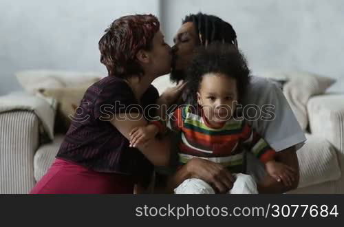 Beautiful mixed couple in loving embrace and sensual kiss. Loving mixed race parents with adorable mixed race child sitting on the floor and enjoying weekend at home. Caucasian mother with eyes closed kissing her african american husband with passion.