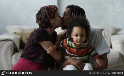 Beautiful mixed couple in loving embrace and sensual kiss. Loving mixed race parents with adorable mixed race child sitting on the floor and enjoying weekend at home. Caucasian mother with eyes closed kissing her african american husband with passion.