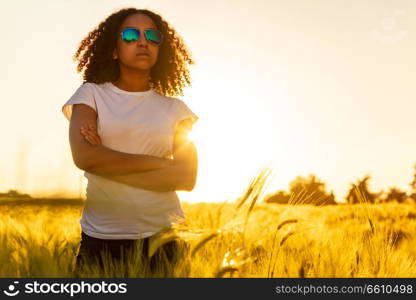 Beautiful mixed race African American female girl teenager young woman wearing sunglassesat sunset or sunrise in a field of wheat, corn or barley