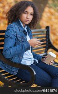Beautiful mixed race African American girl teenager female young woman using cell phone and drinking takeaway coffee outside sitting on a park bench in autumn or fall looking sad depressed or thoughtful