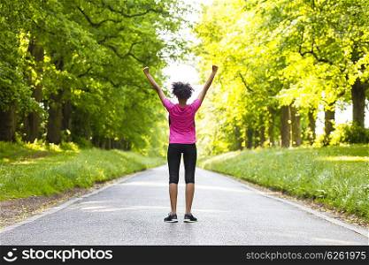 Beautiful mixed race African American young woman girl teenager fitness running jogging celebrating success on road lined with spring or summer green trees