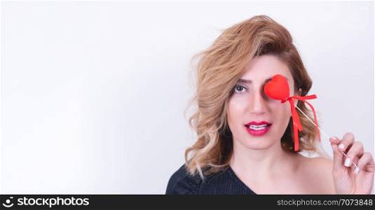 Beautiful model Girl holds Valentine Heart shaped stick over her eyes.Attractive woman holding red love heart stick isolated on white background.