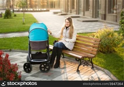 Beautiful mother sitting on bench at park and swaying baby stroller