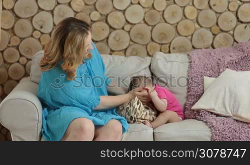 Beautiful mother sitting on sofa and holding her infant child on lap while relaxing at home. Adorable baby girl holding mom&acute;s finger while sitting on her lap. Charming mommy cuddling and nursing her child in living room.