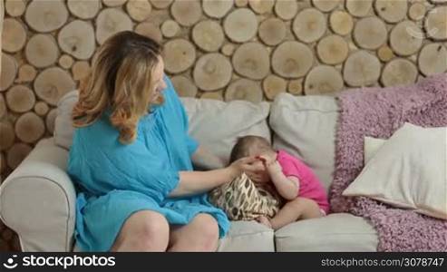 Beautiful mother sitting on sofa and holding her infant child on lap while relaxing at home. Adorable baby girl holding mom&acute;s finger while sitting on her lap. Charming mommy cuddling and nursing her child in living room.