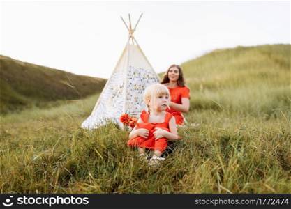 Beautiful mother with her little daughter sitting near wigwam in the field. Spending time together, outside, on vacation, outdoors. Beautiful sunset light in the garden or in the park.. Beautiful mother with her little daughter sitting near wigwam in the field. Spending time together, outside, on vacation, outdoors. Beautiful sunset light in the garden or in the park