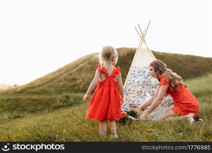 Beautiful mother with her little daughter sitting near wigwam in the field. Spending time together, outside, on vacation, outdoors. Beautiful sunset light in the garden or in the park.. Beautiful mother with her little daughter sitting near wigwam in the field. Spending time together, outside, on vacation, outdoors. Beautiful sunset light in the garden or in the park