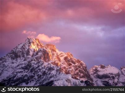 Beautiful mountain peaks in snow and violet sky with pink clouds in winter at sunset. Colorful landscape with high snowy rocks, cloudy purple sky in cold evening. Alpine mountains. Nature background