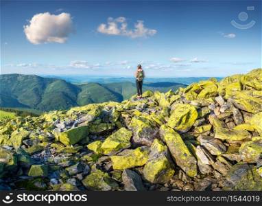 Beautiful mountains and standing young woman with backpack on the green stones at sunset in summer. Landscape with sporty girl on the mountain peak, forest, hills , blue sky with clouds. Travel