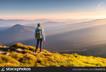 Beautiful mountains in fog and standing young woman with backpack on the peak at sunset in summer. Landscape with sporty girl, green grass, forest, hills , blue sky with sunbeams. Travel and tourism