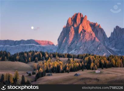 Beautiful mountains with lighted peaks at sunset. Autumn landscape with small wooden houses, mountain valley, meadows with green grass, fall trees, high rocks, sky with moon. Alpe di Siusi in Italy