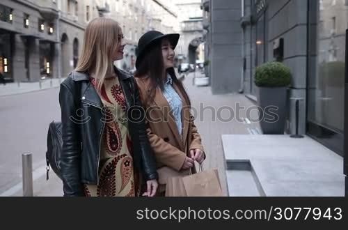 Beautiful multiethnic girlfriends with shopping bags looking in boutique display window outdoors. Lovely young shopaholic females looking for clothes, excited pointing the store window. Happy teenage girls shopping in trade centre.