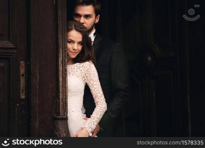 Beautiful newlyweds hugging near the ancient door. Wedding portrait of a stylish groom and a young bride near old house in in a European town.. Beautiful newlyweds hugging near the ancient door. Wedding portrait of a stylish groom and a young bride near old house in in a European town
