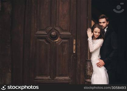 Beautiful newlyweds hugging near the ancient door. Wedding portrait of a stylish groom and a young bride near old house in in a European town.. Beautiful newlyweds hugging near the ancient door. Wedding portrait of a stylish groom and a young bride near old house in in a European town