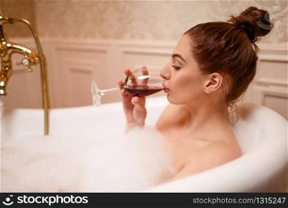 Beautiful nude woman drinking red wine in the bathtub with foam.