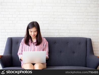 Beautiful of portrait asian young woman working online laptop sitting on sofa at living room, girl using notebook computer with connect to internet for distance job, business and success concept.