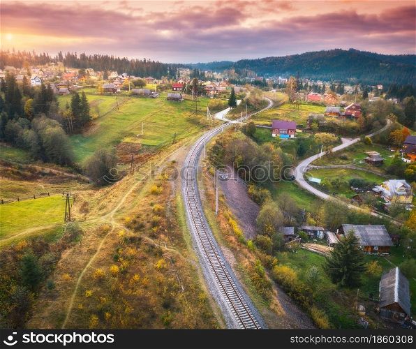 Beautiful old railway station and village at sunset in carpathian mountains in autumn in Ukraine. Aerial view of railroad, green meadows, trees, houses, hills and colorful red sky in fall. Top view