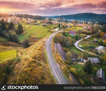 Beautiful old railway station and village at sunset in carpathian mountains in autumn in Ukraine. Aerial view of railroad, green meadows, trees, houses, hills and colorful red sky in fall. Top view