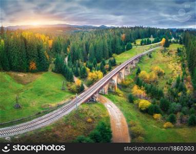 Beautiful old viaduct at sunset in carpathian mountains in autumn in Ukraine. Aerial view of railway bridge, railroad, rural dirt road, green meadows, trees, hills and cloudy sky in fall. Top view
