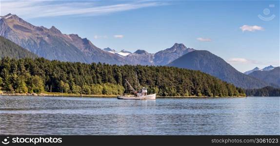 Beautiful panoramic shot of a commercial fishing boat anchored in a harbour in Sitka, AK. Green forest, mountains with snowy peaks, and gorgeous blue sky in the background.