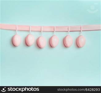 Beautiful pastel pink Easter eggs hanging on ribbon at light at blue turquoise background. Front view, copy space for greeting or invitation