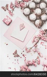 Beautiful pastel pink Easter greeting card mock up with blossom decoration, hearts, eggs in carton box on white desk background, top view, flat lay.