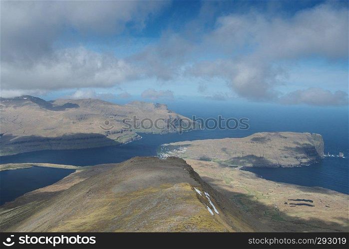 Beautiful perspective scenic view to the landscape with eroded mountains in green and snow on hillsides and village with ocean in background on a sunny day with rich clods over the Faroe Islands for best travel backgrounds. Glorious sceneries of the Faroes. Postcard motif.