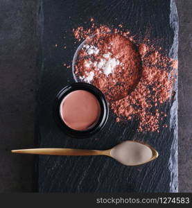 Beautiful photo of pink clay skincare product in open elegant black glass jar near golden spoon filled with creamy white clay surrounded by dry clay sprinkles on black stone plate surface