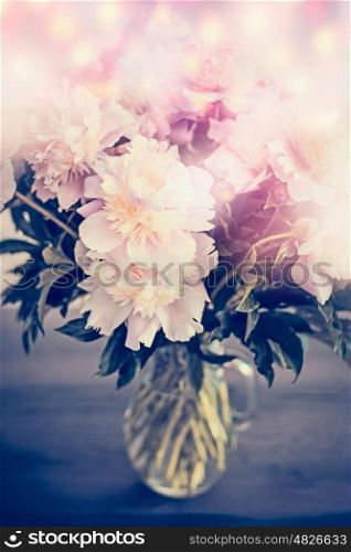 Beautiful pink pale peonies bunch in glass vase on table with bokeh lighting. Romantic flowers bouquet, front view