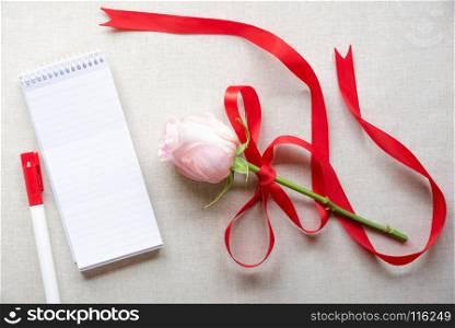 Beautiful pink rose tied with red satin ribbon with bow, an unwritten spiral notebook with stripes and a marker pen, on a vintage fabric background.