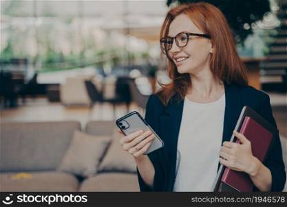 Beautiful pleased redhead businesswoman in formal outfit holding mobile phone in one hand and laptop with notebook in another and smiling while looking aside, waiting for business partner in office. Beautiful pleased redhead businesswoman in formal outfit holding mobile phone in one hand