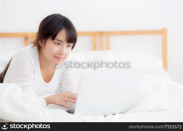 Beautiful portrait asian young woman smile lying on bed using laptop computer with happy at bedroom for leisure and relax, freelance with girl working notebook, communication concept.