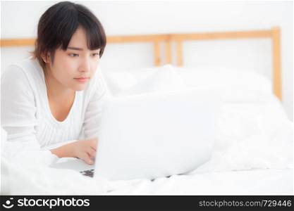 Beautiful portrait asian young woman smile lying on bed using laptop computer with happy at bedroom for leisure and relax, freelance with girl working notebook, communication concept.