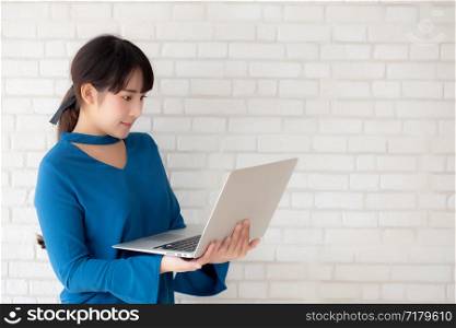 Beautiful portrait asian young woman smile using laptop standing at workplace on cement concrete background, girl happy with computer internet online, lifestyle and freelance business concept.